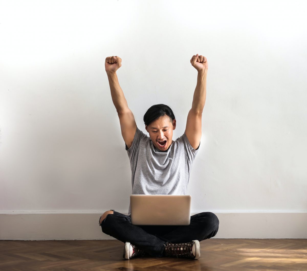 A young man with black hair and light brown skin raises his hands in triumph over his laptop.