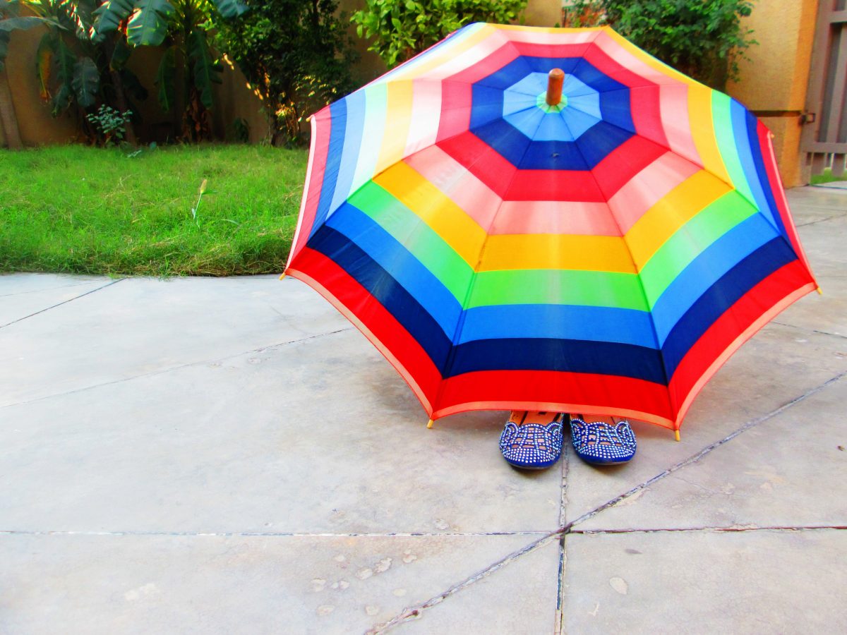 A pair of blue shoes peek out from underneath a multi-colored umbrella. 