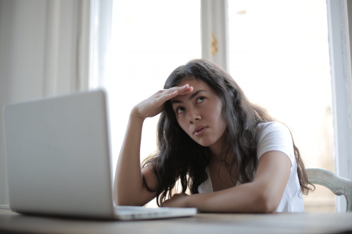 A young woman with long, wavy brown hair sits in front of a laptop with a hand on her forehead and an irritated look on her face.